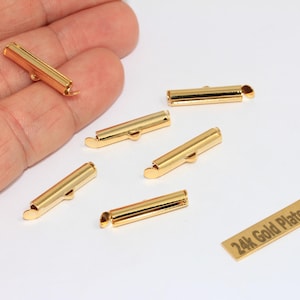 Puede incluir: Cinco terminales de metal de color dorado para joyería, cada uno con forma cilíndrica y una pequeña anilla de sujeción. Una etiqueta rectangular con el texto 24k Gold Plated se encuentra cerca de los componentes metálicos.