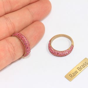 Puede incluir: Dos anillos dorados con piedras rosas. Un anillo está en un dedo, el otro está separado. También hay una pequeña etiqueta rectangular de latón con la inscripción "Raw Brass".