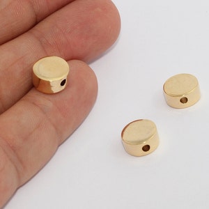 May include: Gold-colored, cylindrical beads with a flat top and bottom. Each bead has a small hole drilled through the center. One bead is held in a hand, with two others scattered nearby on a white surface. These beads are likely for jewelry making.