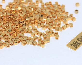 May include: A collection of small, gold-colored, square beads with rounded edges. The beads are scattered on a white surface. A rectangular tag with the text "24k Gold Plated" is also present.