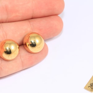 May include: Two shiny, gold-colored, round stud earrings. The earrings are smooth and reflective, catching the light. A small, rectangular tag with the text "24k Gold Plated" is also visible.