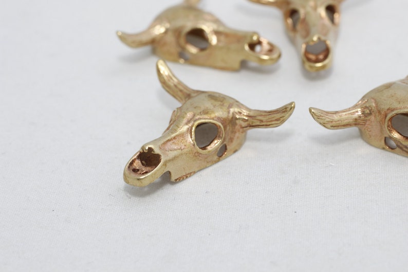 May include: Close-up of several small, gold-colored bull skull charms. Each charm has detailed horns, eye sockets, and a nasal cavity. The charms are arranged on a white surface, suggesting a craft or jewelry-making theme.
