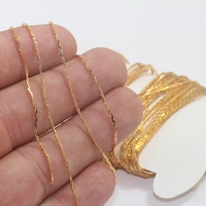 Puede incluir: Primer plano de una cadena dorada con un diseño retorcido y texturizado. La cadena se sostiene sobre un fondo blanco, mostrando sus intrincados detalles y superficie reflectante. Ideal para la fabricación de joyas o manualidades.