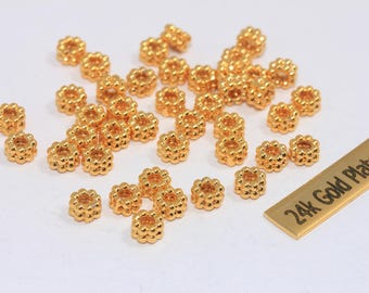 May include: A collection of small, gold-colored beads with a floral design. The beads are scattered on a white surface, with a small rectangular tag that reads "24k Gold Plated". These beads are likely used for jewelry making or other craft projects.