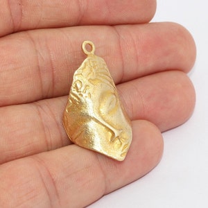 Może przedstawiać: Złoty wisiorek z rzeźbionym wzorem twarzy. Wisiorek ma pętlę u góry do zawieszenia i teksturowaną powierzchnię. Twarz jest w profilu, ze szczegółowymi cechami. Wisiorek jest trzymany w dłoni.