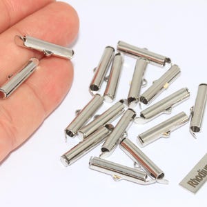 May include: A collection of silver-colored, cylindrical jewelry clasps with small loops. The clasps are scattered on a white surface, with some held in a hand. A small metal tag reads "Rhodium Plated."
