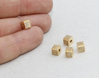 May include: Gold-colored cubic beads with embedded clear crystals. The beads are small, with a square shape and a hole running through the center. The crystals are arranged in a grid pattern on each face of the cube.