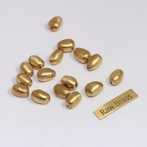 Może przedstawiać: Kolekcja złotych, owalnych koralików rozrzuconych na białej powierzchni. Obecna jest również mała, prostokątna mosiężna etykieta z napisem "Raw Brass". Koraliki te są prawdopodobnie przeznaczone do tworzenia biżuterii lub rękodzieła.