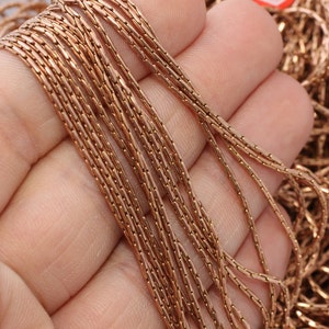 Puede incluir: Primer plano de un montón de cadenas de color cobre. Varios hilos de la cadena están enfocados, revelando un patrón detallado y texturizado. La cadena está hecha de pequeños eslabones rectangulares, creando un diseño delicado e intrincado. Las cadenas se sostienen en una mano.