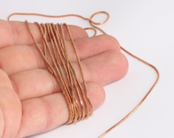 May include: A coiled length of copper-colored snake chain, likely for jewelry making. The chain is held in a hand against a white background. The chain has a smooth, reflective surface.