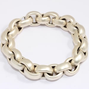 Puede incluir: Un brazalete de cadena plateada con eslabones grandes y redondeados. El brazalete está hecho de un metal mate y se muestra sobre un fondo blanco. Los eslabones están entrelazados, formando una forma circular.