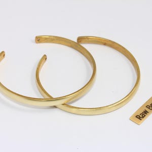 Op de afbeelding: Twee goudkleurige messing armbanden met een glad, gebogen ontwerp. De armbanden zijn open en liggen op een wit oppervlak. Een klein rechthoekig label met de tekst "Raw Brass" is ook aanwezig.