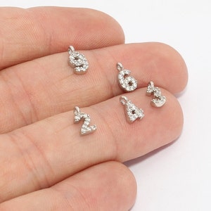 May include: A collection of small, silver-colored number charms, each encrusted with small, clear crystals. The numbers 2, 3, 4, 6, and 9 are visible. Each charm has a small loop at the top for attaching to a chain or bracelet. These charms are ideal for jewelry making.