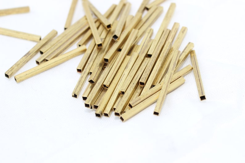 May include: A collection of small, rectangular brass tubes with square openings at each end. The tubes are scattered on a white surface, with some overlapping. The brass has a warm, golden color.