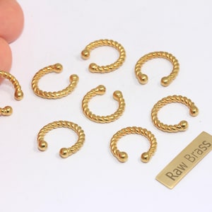 May include: Close-up of several gold-colored, C-shaped ear cuffs with a twisted rope design. A small rectangular tag with the text "Raw Brass" is also visible. The ear cuffs are arranged on a white surface.