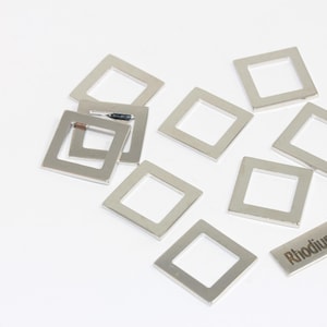 Puede incluir: Un conjunto de componentes metálicos cuadrados con baño de rodio, dispersos sobre una superficie blanca. Una pieza rectangular está grabada con el texto "Rhodium Plated". Los componentes tienen un aspecto plateado brillante.