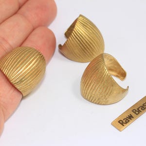 May include: Three raw brass rings with a ridged design, one worn on a finger. A small brass tag with the text "Raw Brass" is also present. The rings have a gold color.