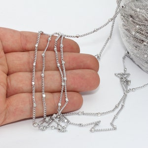 Könnte beinhalten: Silberfarbene Kette mit kleinen, gleichmäßig verteilten Perlen. Die Kette ist über einer Hand drapiert, wobei eine Spule derselben Kette im Hintergrund sichtbar ist. Die Kette hat ein zartes, glänzendes Aussehen, geeignet für die Schmuckherstellung.