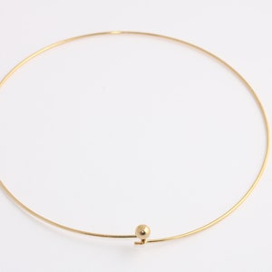 May include: A gold-colored choker necklace with a delicate, minimalist design. The necklace features a thin, circular band with a small, spherical accent near the clasp. The jewelry is set against a plain white background.