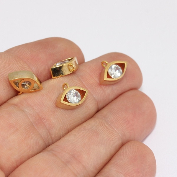 Op de afbeelding: Goudkleurige, oogvormige bedels met een heldere, ronde kristal in het midden. De bedels hebben een kleine lus aan de bovenkant om aan sieraden te bevestigen. De bedels worden tegen een lichte achtergrond getoond.