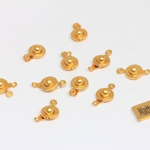 May include: A collection of gold-colored jewelry clasps with a round, domed center and loop attachments. A small rectangular tag with the text "Matt Gold Plated" is also present. These clasps are likely used for necklaces or bracelets.