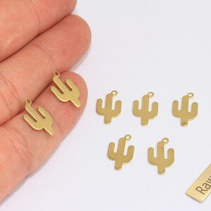 Könnte beinhalten: Goldfarbene Kaktus-Anhänger mit einer kleinen Schlaufe oben zur Schmuckherstellung. Die Anhänger sind auf einer weißen Oberfläche angeordnet. Ein kleines rechteckiges Etikett mit dem Text "Raw Brass" ist ebenfalls vorhanden.