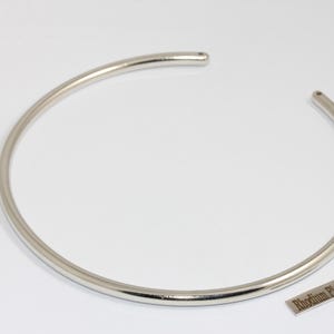 May include: A silver-colored, open-ended collar necklace. The necklace is a simple, smooth, and rounded design. A small rectangular tag with the text "Rhodium Plated" is next to the necklace.