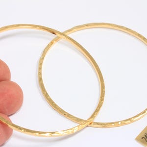 May include: Two gold-plated hoop earrings with a hammered texture. The earrings are circular and have a slightly irregular surface. A small tag reads "24k Gold Plated". The earrings are shown against a white background.