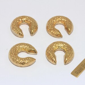 May include: Four gold-colored, crescent-shaped jewelry pieces with a textured, hammered surface. A small, rectangular brass tag with the text "Raw Brass" is also present. The pieces are arranged on a white background.