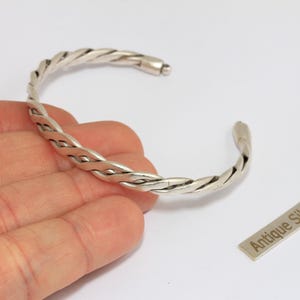 Può includere: Un bracciale a polsino intrecciato color argento. Il bracciale è aperto e presenta un design intrecciato. È presente anche una piccola etichetta rettangolare con la scritta "Antique Silver".