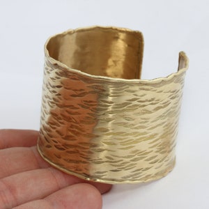 May include: A wide, gold-coloured cuff bracelet with a textured surface. The bracelet has a hammered appearance and is open on one side. The cuff is approximately 5 cm wide.