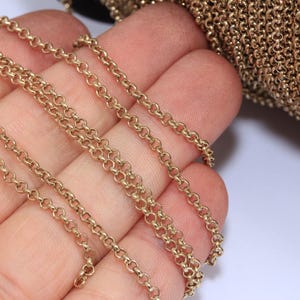 May include: Close-up of a gold-colored chain with small, circular links. The chain is held in a hand, with a portion of a larger roll of chain visible in the background. The chain has a shiny, metallic appearance.