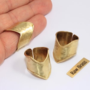 May include: Three hammered brass rings and a small brass rectangle with the text "Raw Brass" are displayed on a white surface. One ring is on a finger, and the other two are next to the rectangle.