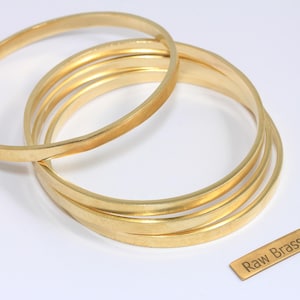 Könnte beinhalten: Eine Sammlung goldfarbener Messingarmreifen. Das Bild zeigt mehrere runde, dünne Armbänder, die übereinander gestapelt sind, mit einem kleinen rechteckigen Etikett mit der Aufschrift "Raw Brass". Die Armreifen werden auf einer weißen Oberfläche präsentiert.