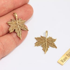 Puede incluir: Dos colgantes dorados con forma de hoja de arce con detalles intrincados. Uno de los colgantes se sostiene en una mano, mientras que el otro está sobre una superficie blanca. También hay una pequeña etiqueta rectangular con el texto "Raw Brass".