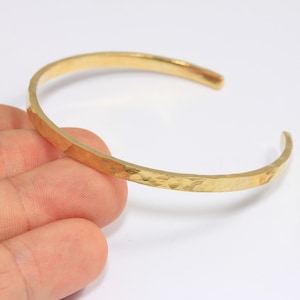 May include: A hammered gold-tone brass cuff bracelet. The open-ended bracelet has a textured surface and a simple, elegant design. A small brass tag with the text "Raw Brass" is also visible.