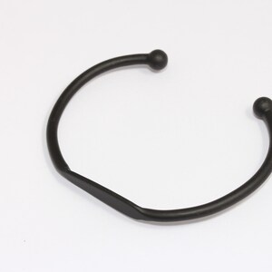 May include: A black metal cuff bracelet with a simple, open design. The bracelet has a curved shape with a small ball at each end. The metal has a matte finish, and the bracelet is set against a white background.