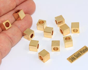 May include: A collection of small, gold-colored, cube-shaped beads with a hole through the center. The beads are scattered on a white surface, with a small rectangular tag that reads "24k Gold Plated".