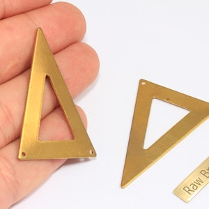 May include: Gold-colored brass triangle charms with a cutout center and small holes for jewelry making. One triangle is held in a hand, and two others are on a white surface. A small brass rectangle with the text "Raw Brass" is also present.