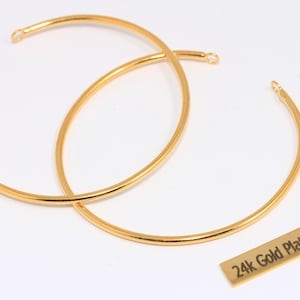 Puede incluir: Dos componentes de joyería de metal dorado y curvado con pequeños bucles en cada extremo, superpuestos sobre un fondo blanco. También hay una pequeña etiqueta rectangular con el texto "24k Gold Plated".