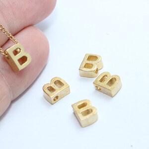 Peut inclure: Un collier avec un pendentif en forme de lettre "B" de couleur or et plusieurs breloques en forme de lettre "B". Le pendentif est sur une chaîne délicate. Les breloques sont petites, carrées et percées. Le fond est blanc.