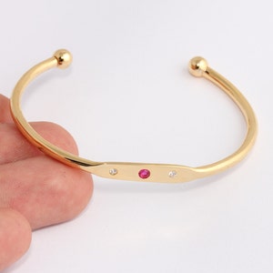 Può includere: Bracciale a polsino color oro con un design rettangolare centrale. Il bracciale presenta una vibrante gemma rossa affiancata da due gemme chiare più piccole. Le estremità del bracciale hanno piccoli accenti dorati rotondi.