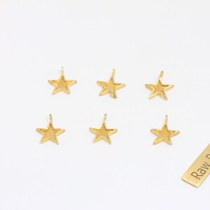 May include: Six gold-colored star charms with a loop at the top for hanging. A small rectangular tag with the words "Raw Brass" is also present. The charms are on a white background.