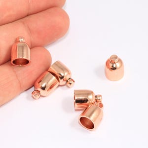 May include: Rose gold-coloured metal bead caps with a small loop and a rounded top. The caps are cylindrical and have a smooth, polished finish. Several caps are shown, some open and some closed.
