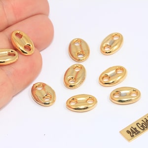 May include: Gold-plated oval link charms with two holes, scattered on a white surface. A small rectangular tag with the text "24k Gold Plated" is also present. These charms are ideal for jewelry making.