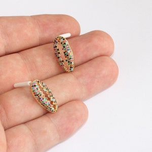 May include: Two gold-colored oval stud earrings. Each earring is adorned with a pattern of colorful gemstones, including red, blue, green, and black. The earrings have white posts for insertion into the ear.
