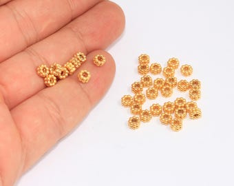 May include: Gold-colored metal beads with a floral design. Some beads are held in a hand, while others are scattered on a white surface. These small beads are suitable for jewelry making and crafting projects.