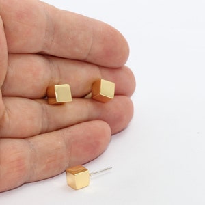 Può includere: Orecchini a lobo a forma di cubo color oro. Gli orecchini sono piccoli, geometrici e hanno un design minimalista. Gli orecchini sono mostrati su sfondo bianco.