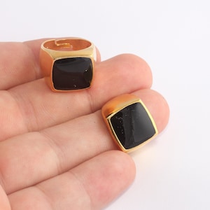 Puede incluir: Dos anillos dorados con una piedra cuadrada de ónix negro. Un anillo está en un dedo, el otro está separado. Los anillos tienen un diseño clásico y audaz. Los anillos probablemente están hechos de metal chapado en oro.