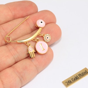 May include: Gold-plated safety pin with pink enamel details and hanging charms. Charms include a Hamsa hand, a pink circle with the letter "I", and an eye-shaped charm. The pin is held in a hand.